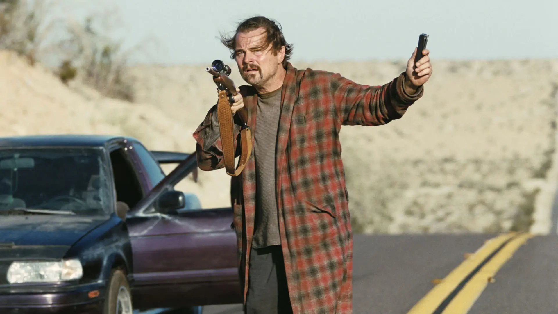 لئوناردو دیکاپریو در میانه‌ی جاده‌ای خارج شهر از ماشین پیاده شده است و اسلحه‌ای دوربین دار در دست دارد در نمایی از فیلم نبرد پشت نبرد پل توماس اندرسون