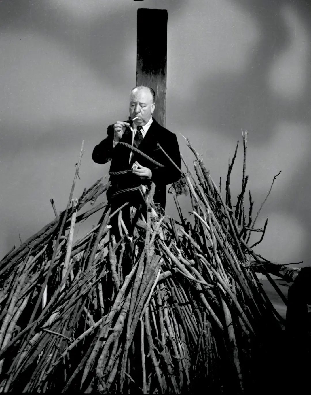 آلفرد هیچکاک (Alfred Hitchcock) در حال کشیدن پیپ روی توده‌ای از شاخه‌ها