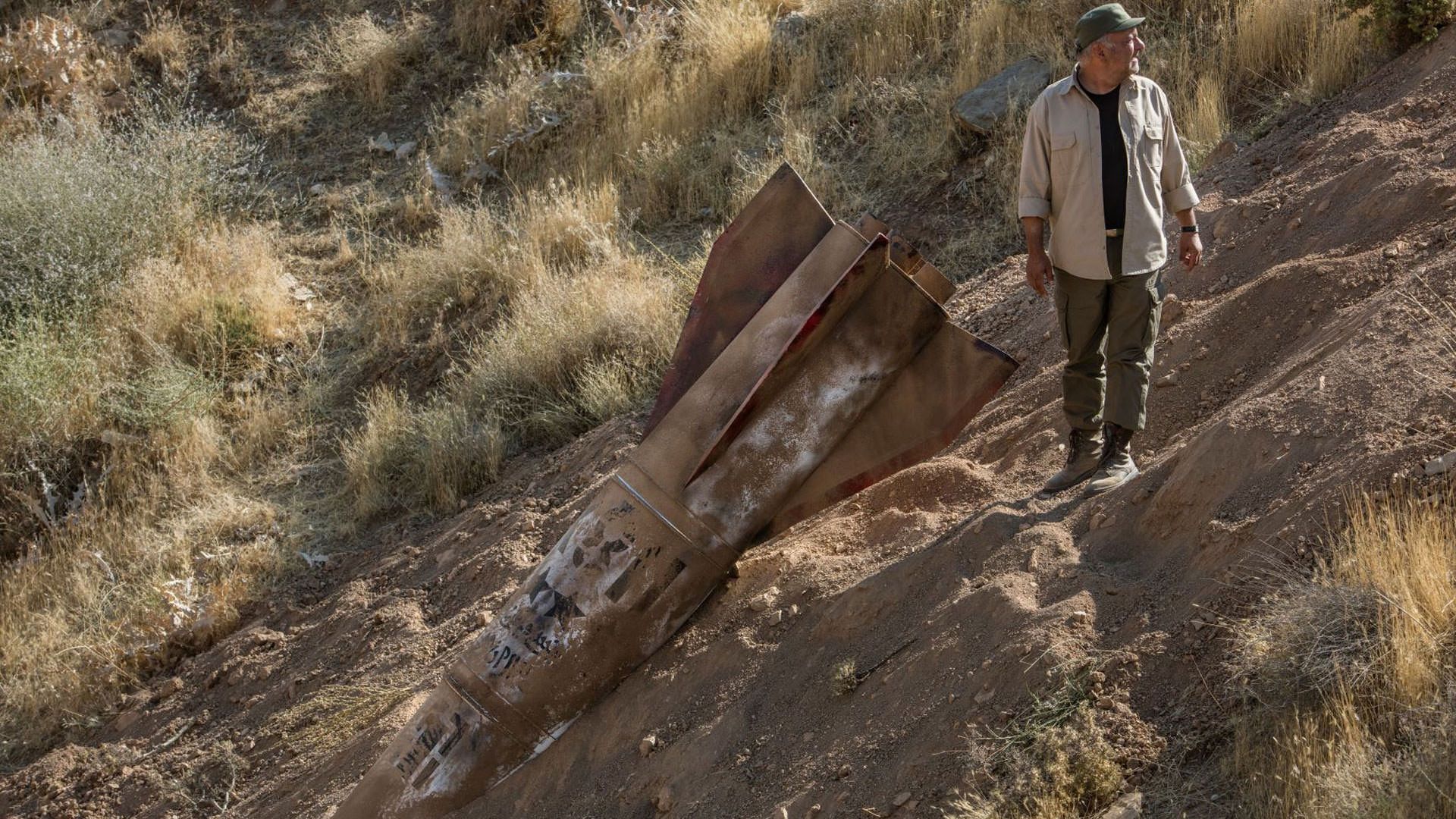 بابک کریمی کنار لاشه‌ی موشکی که در خاک فرو رفته است ایستاده در نمایی از فیلم جنایت بی‌دقت به کارگردانی شهرام مکری