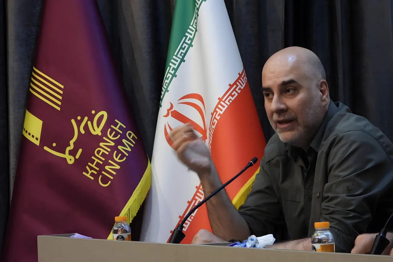 مردی در حال صحبت در نشست سینمایی با پرچم ایران