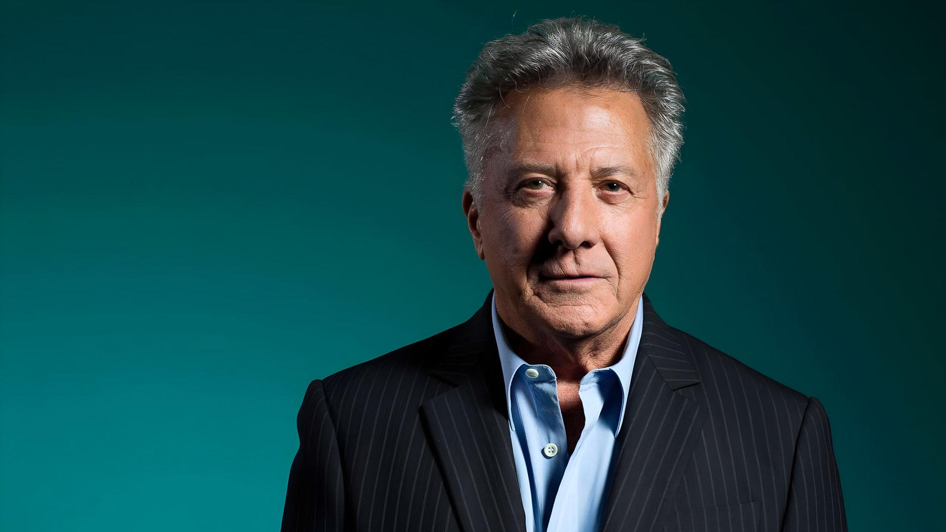 داستین هافمن (Dustin Hoffman) با کت سرمه‌ای و پیراهن آبی در مقابل پس‌زمینه سبز