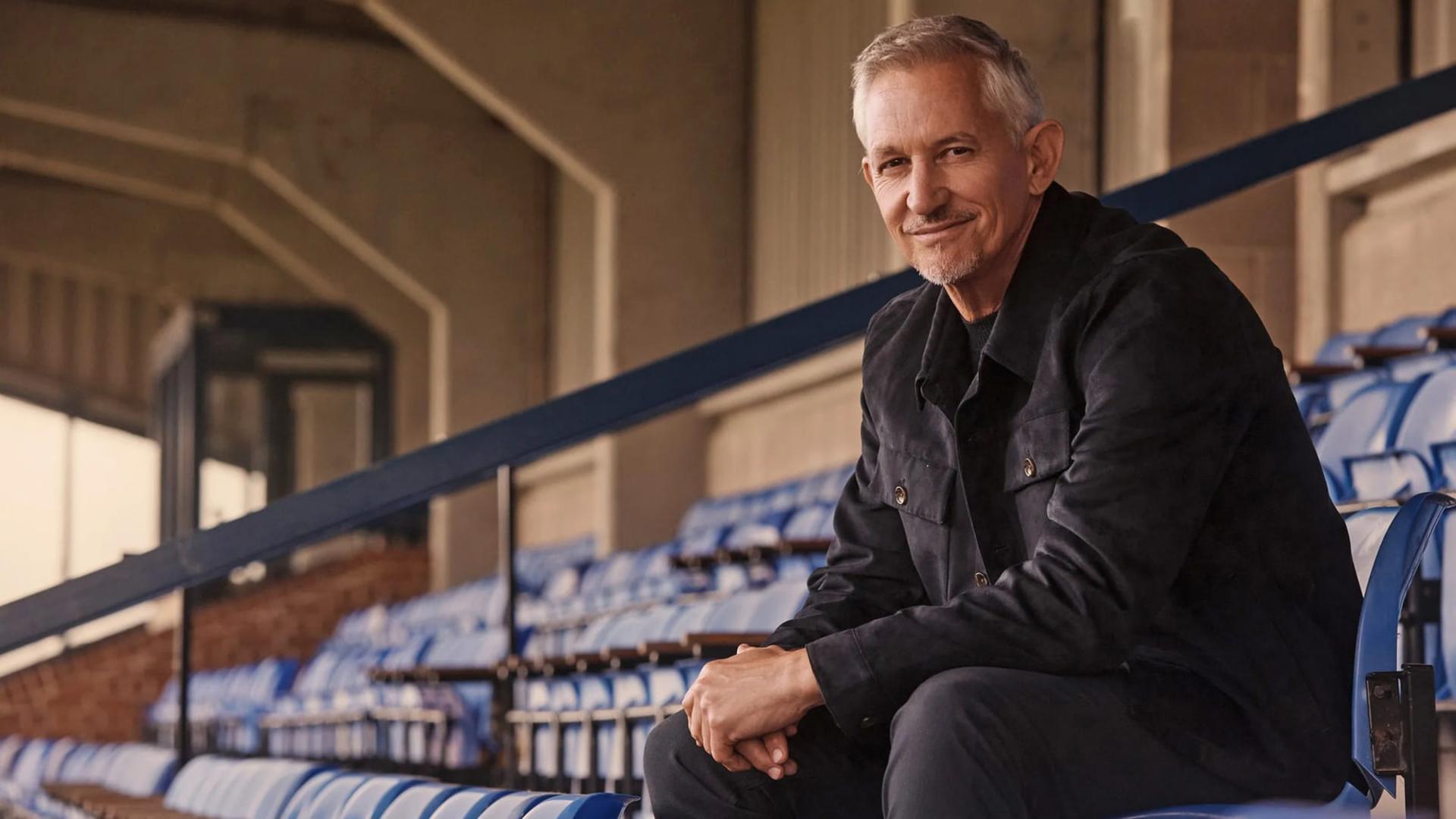 گری لینکر (Gary Lineker) روی صندلی‌های آبی رنگ ورزشگاه نشسته است