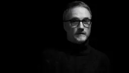 دیوید فینچر (David Fincher) با عینک و لباس تیره در پس‌زمینه سیاه