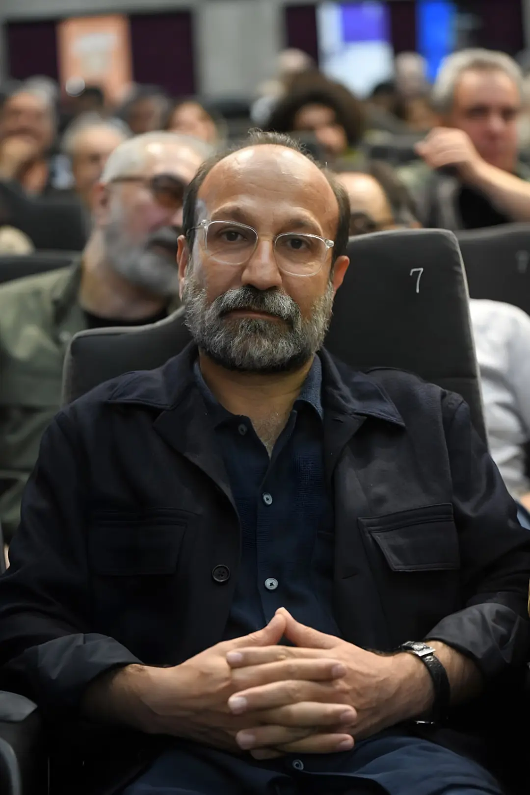 فرهادی (Farhad) با عینک و ریش در سالن مراسم نشسته‌ است