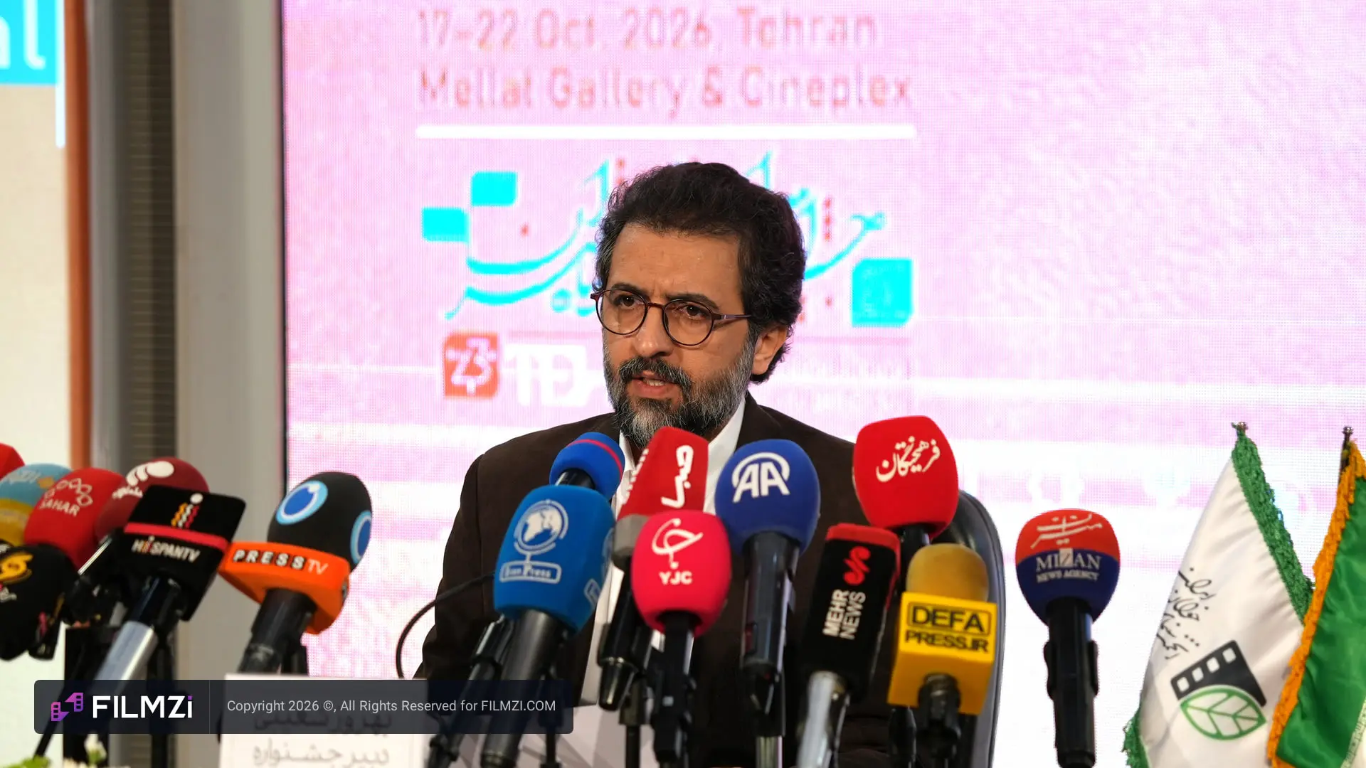 مردی با عینک در میان میکروفون‌های رسانه‌های مختلف در کنفرانس مطبوعاتی