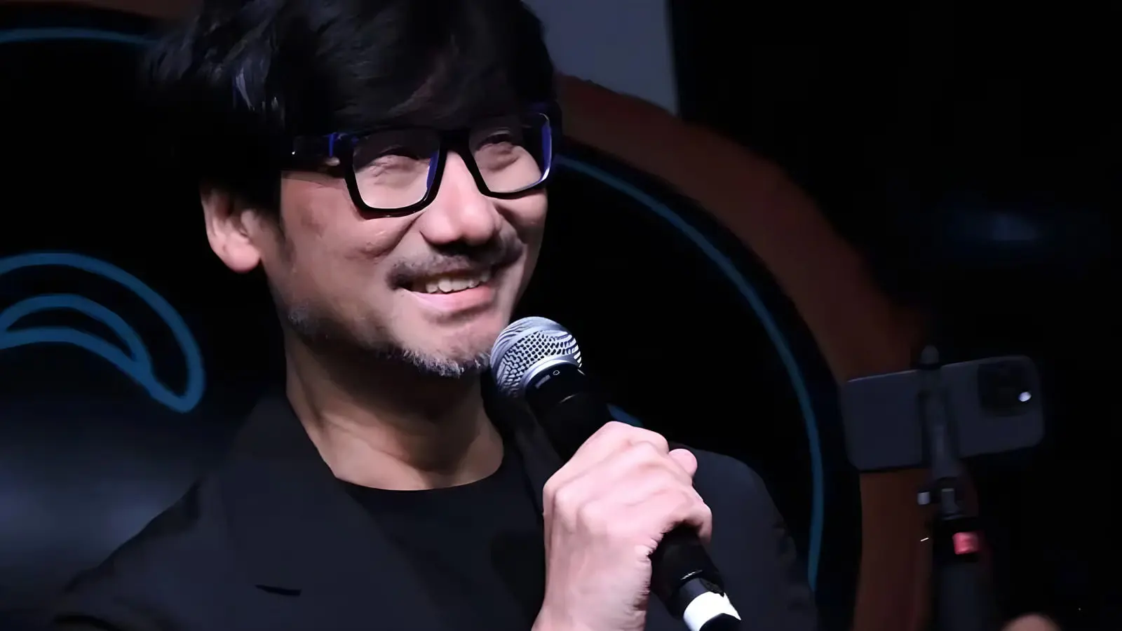 هیدئو کوجیما (Hideo Kojima) با عینک مشکی در حال صحبت با میکروفون