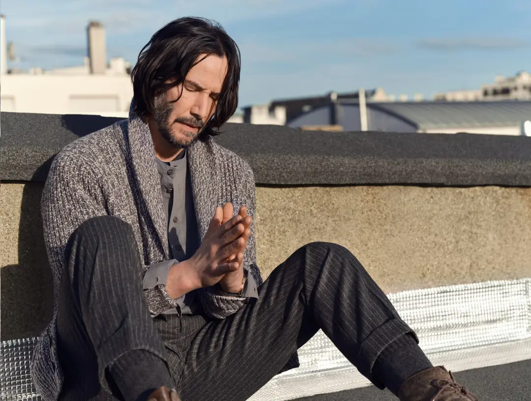 کیانو ریوز (Keanu Reeves) با دست‌های گره‌خورده روی لبه یک پشت‌بام