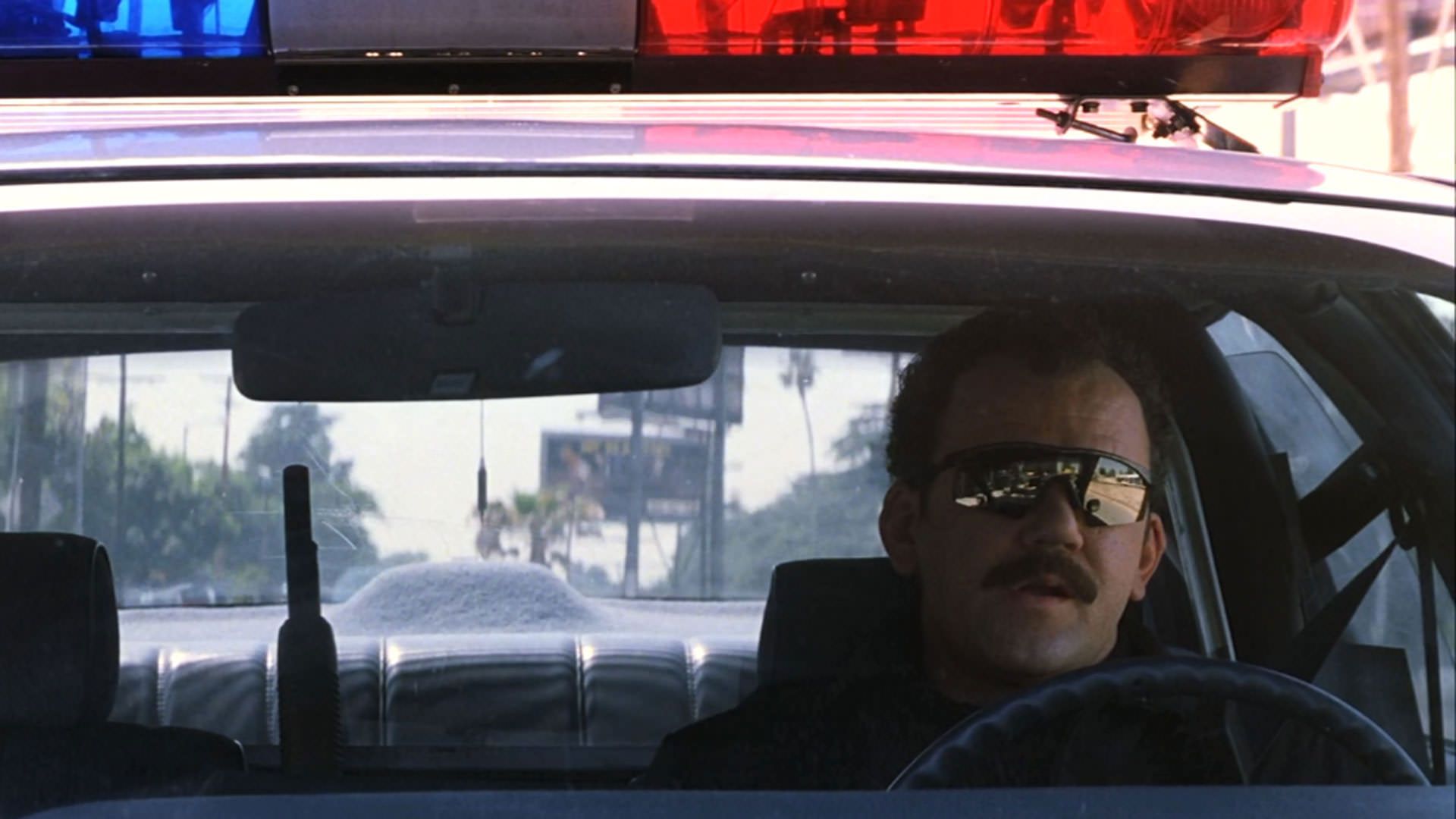 جان سی رایلی با سبیل و لباس پلیس یک ماشین پلیس را میراند در نمایی از فیلم ماگنولیا به کارگردانی پل توماس اندرسون