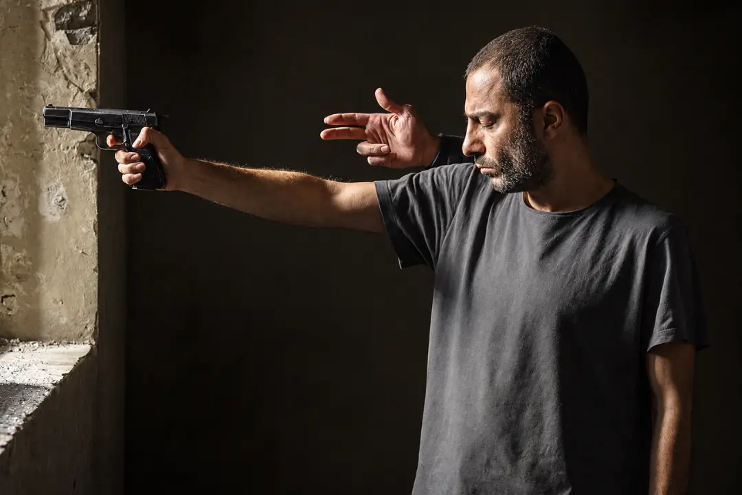 مردی با تشر و نگاهی متمرکز در حال نگه داشتن یک تپانچه در محیطی تاریک