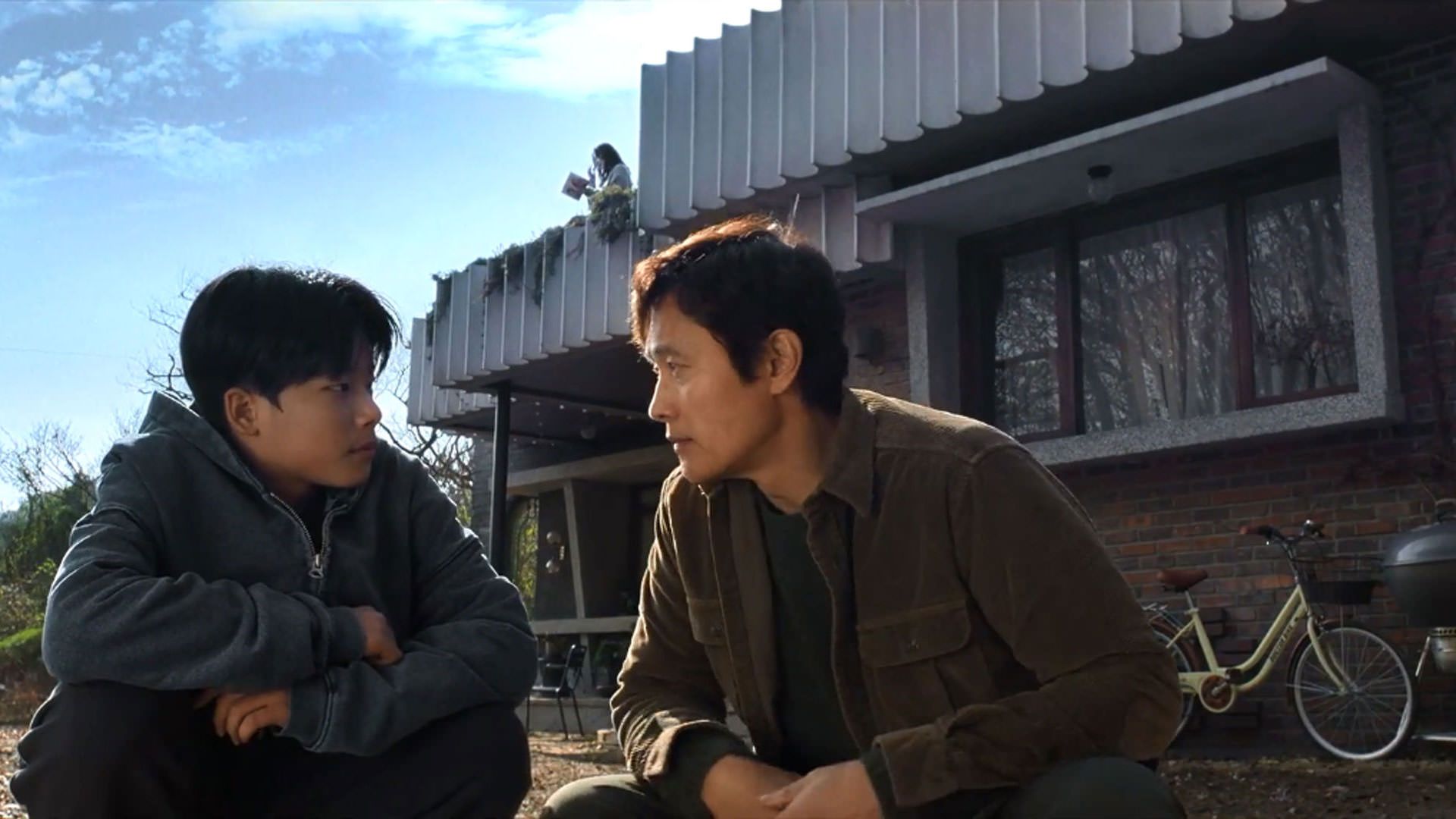 لی بیونگ هون نشسته کنار یک پسر نوجوان پشت به خانه‌ای بزرگ در نمایی از فیلم چاره‌ای نیست به کارگردانی پارک چان ووک