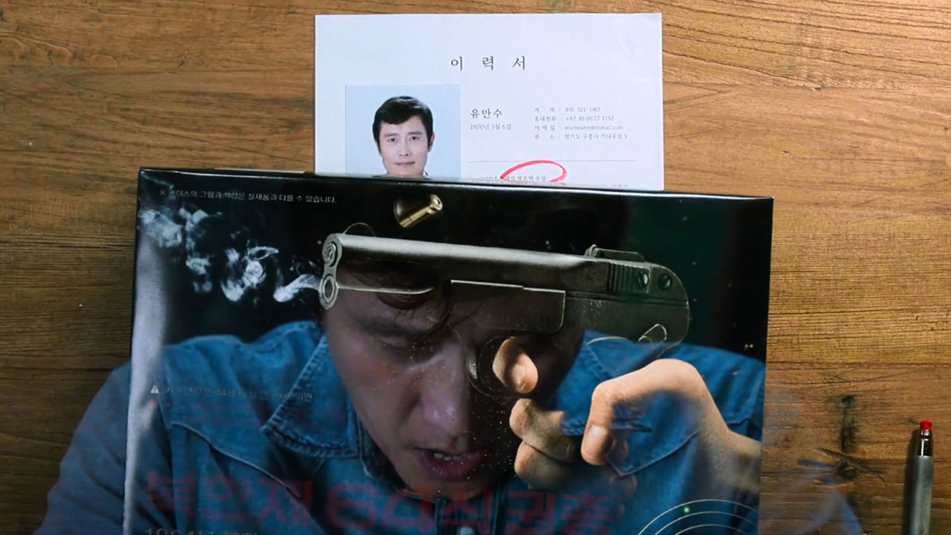 بازتاب تصویرِ لی بیونگ هون روی جعبه‌ی تفنگ در نمایی از فیلم چاره‌ای نیست به کارگردانی پارک چان ووک