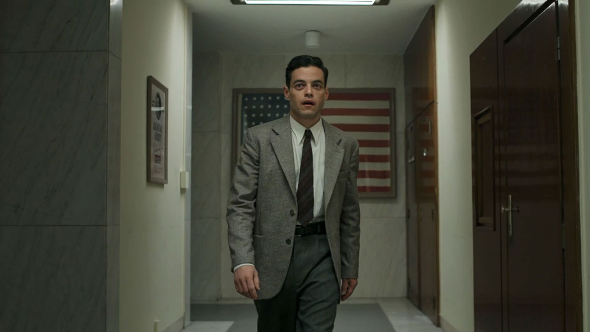 رامی مالک با کت و شلوار پشت به پرچم آمریکا رو به دوربین قدم می‌زند در نمایی از فیلم نورنبرگ جیمز وندربیلت