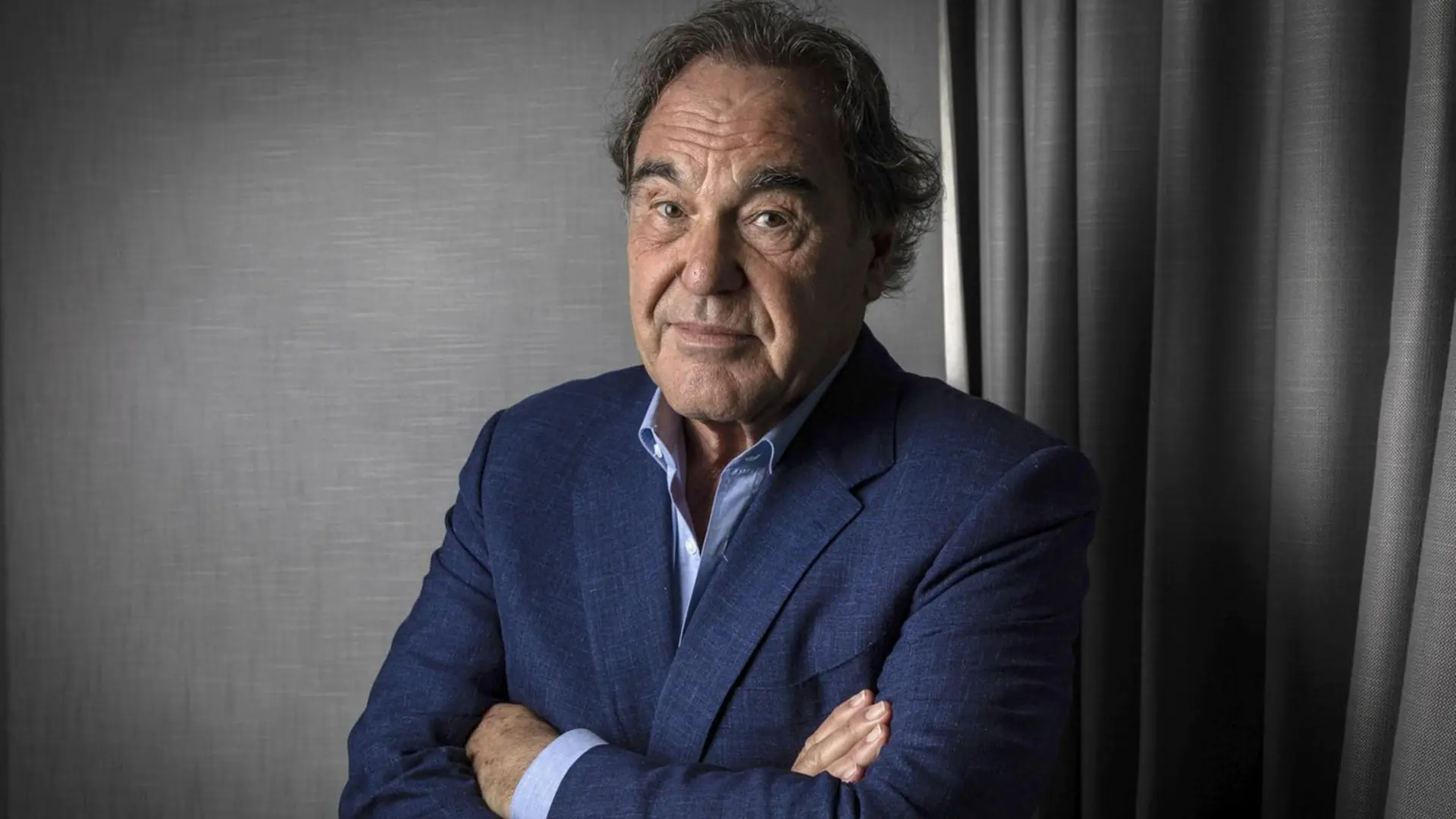 اولیور استون (Oliver Stone) با دست‌های گره‌خورده در مقابل پس‌زمینه خاکستری