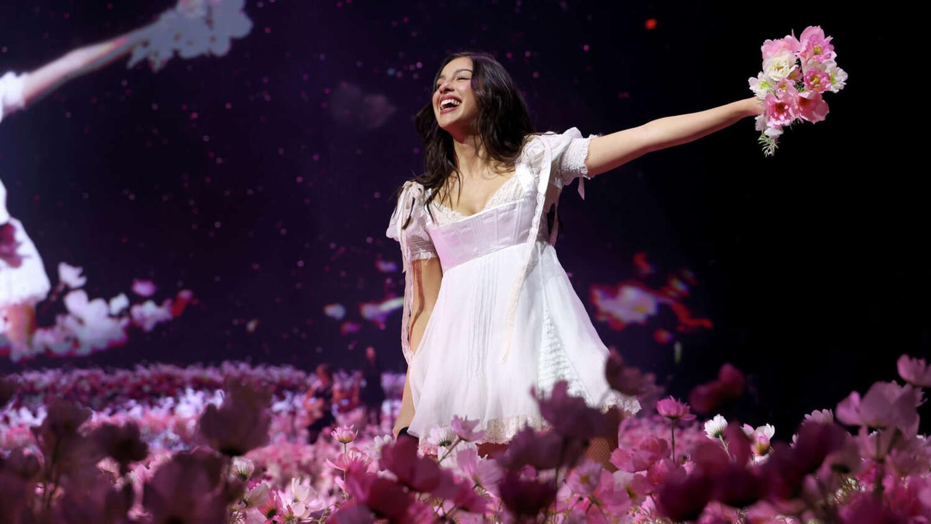 اولیویا رودریگو (Olivia Rodrigo) با لباس سفید در میان گل‌های صورتی روی صحنه