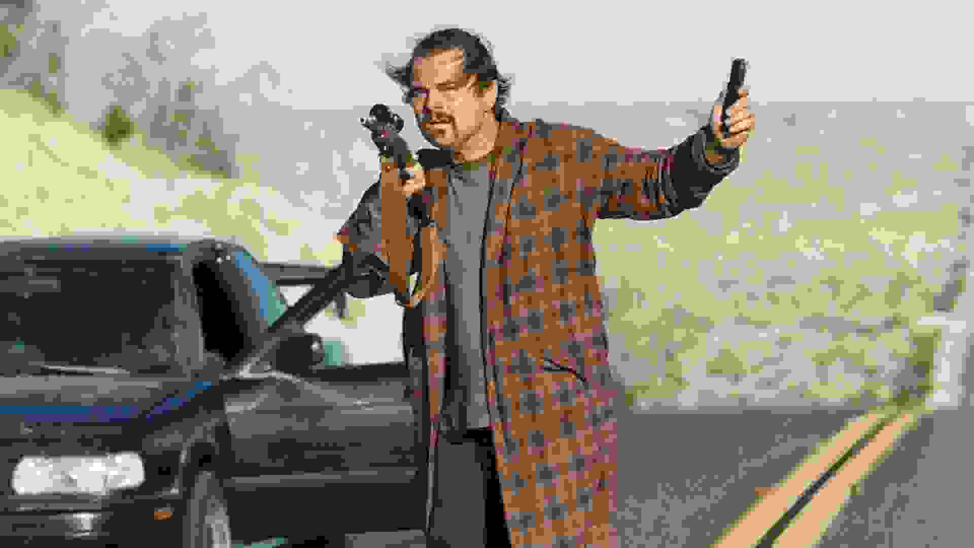 لئوناردو دیکاپریو در میانه‌ی جاده‌ای خارج شهر از ماشین پیاده شده است و اسلحه‌ای دوربین دار در دست دارد در نمایی از فیلم نبرد پشت نبرد پل توماس اندرسون