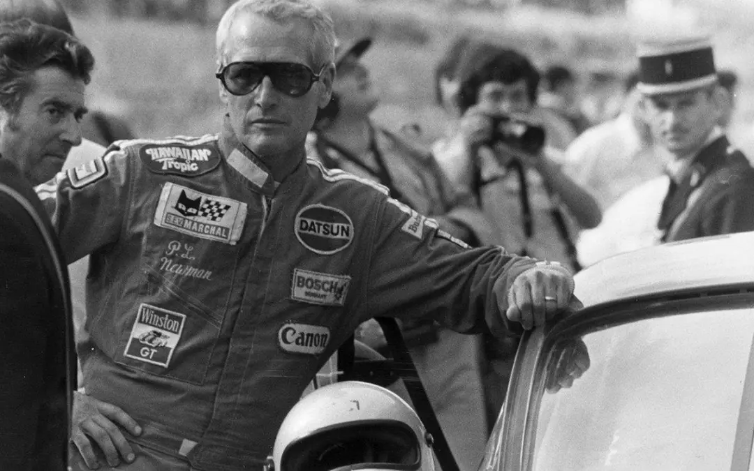 پل نیومن (Paul Newman) با لباس مسابقه‌ای و عینک آفتابی در کنار خودرو