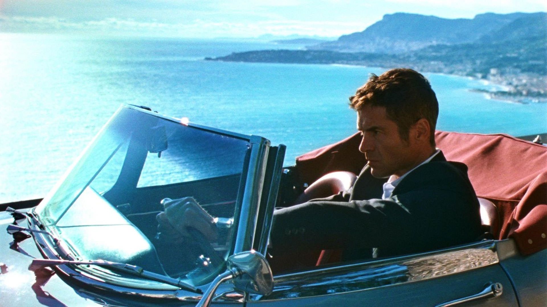 یانیک رنیه در جاده‌ای با منظره‌ی دریا می‌راند در نمایی از فیلم بازتاب در الماس مرده به‌ کارگردانی هلن کاته و برونو فورزانی