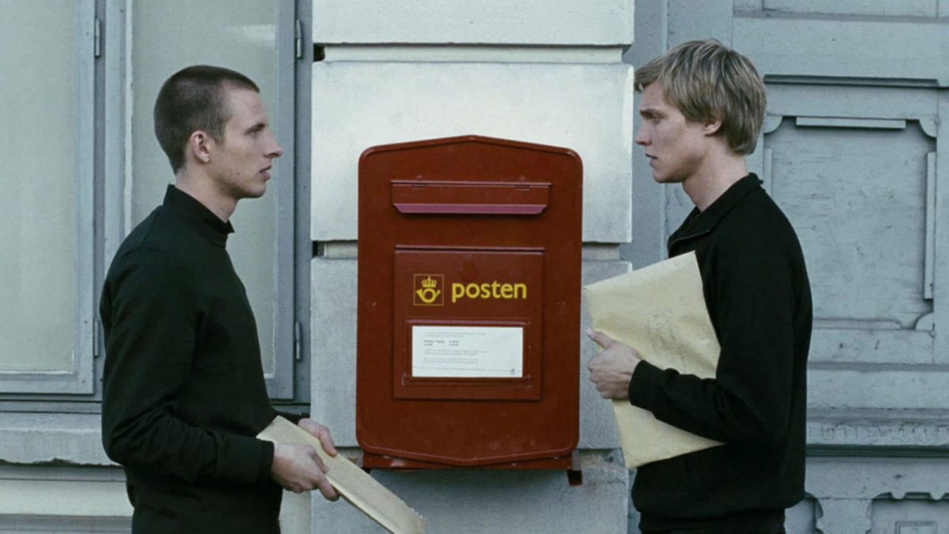 آندرس دنیلسن لی با پیراهنی مشکی ایستاده مقابل اسپن کلومن هوینر کنار صندوق پستی قرمز رنگی که روی دیواری سفید نصب شده است در نمایی از فیلم تکرار به کارگردانی یواکیم تریه