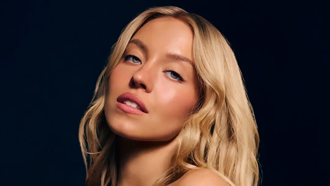 چهره نزدیک سیدنی سوئینی (Sydney Sweeney) با موهای بلوند در پس‌زمینه تیره