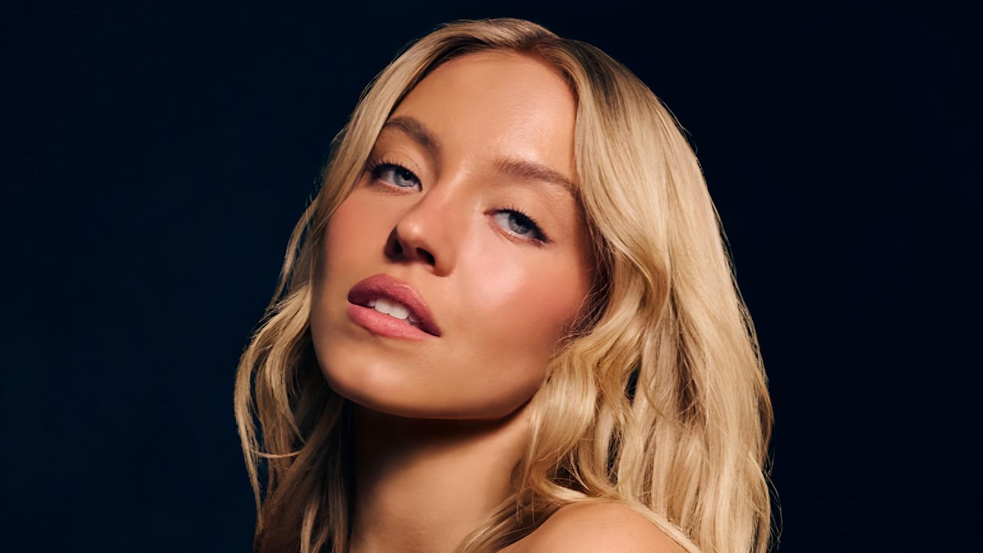 چهره نزدیک سیدنی سوئینی (Sydney Sweeney) با موهای بلوند در پس‌زمینه تیره