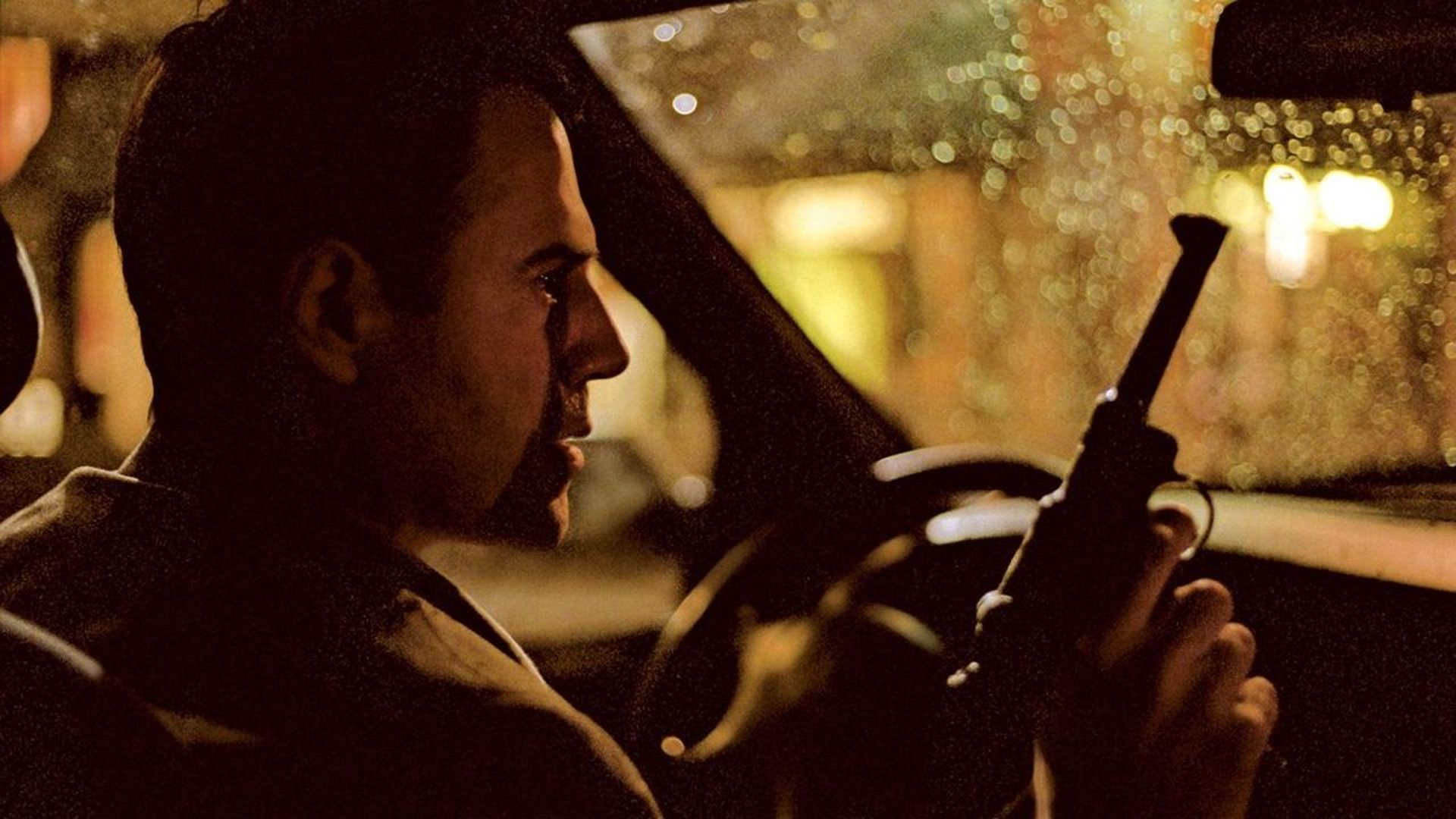 خوزه گارسیا با تفنگی در دست شبانه داخل ماشینی نشسته که نور زرد قطرات باران روی پنجره‌اش را روشن کرده است در نمایی از فیلم تبر به کارگردانی کوستا گاوراس