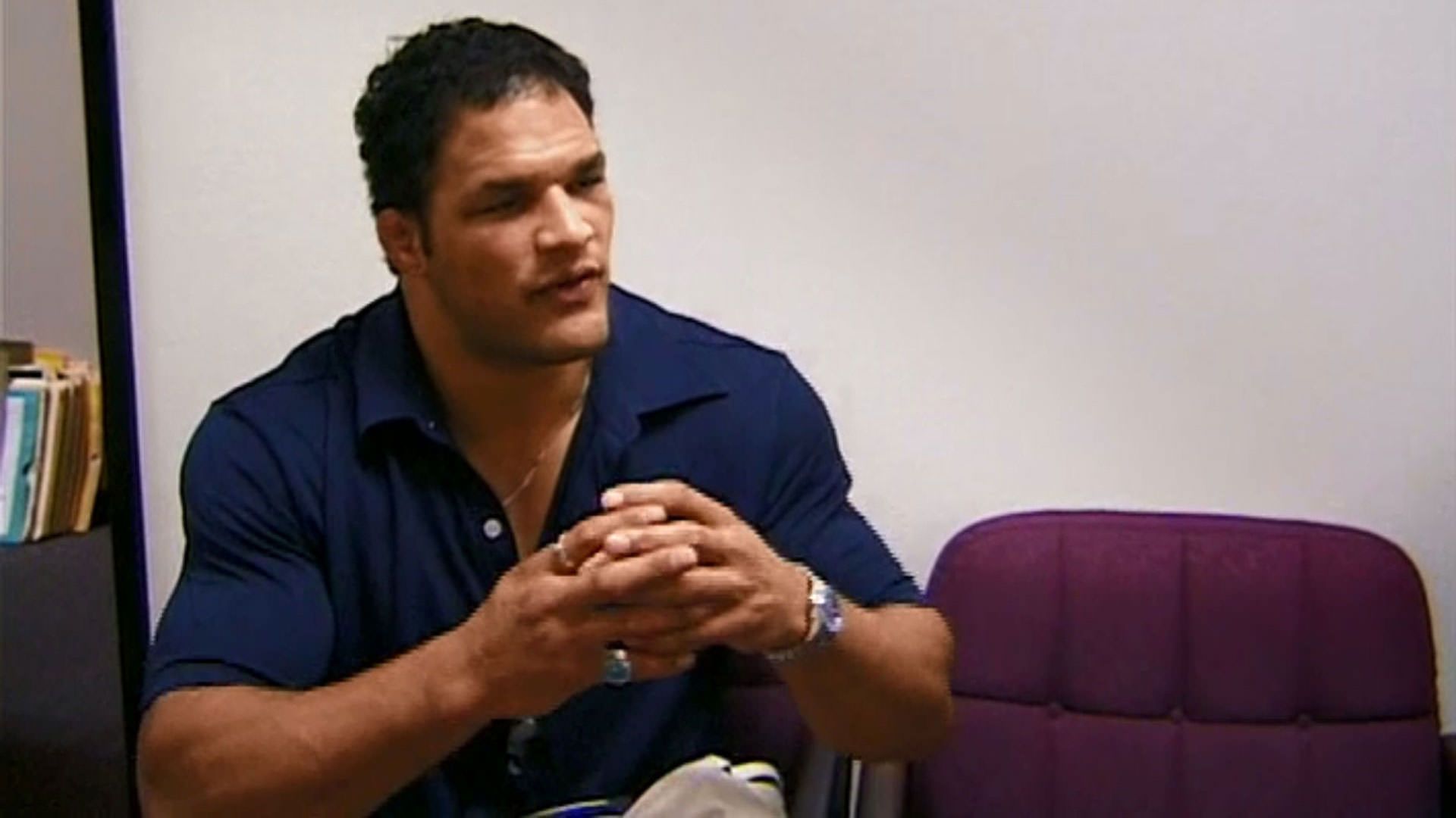 مارک کر با لباس سرمه‌ای روی صندلی نشسته است و چیزی را توضیح می‌دهد در نمایی از مستند ماشین کوبنده به کارگردانی جان هایمز