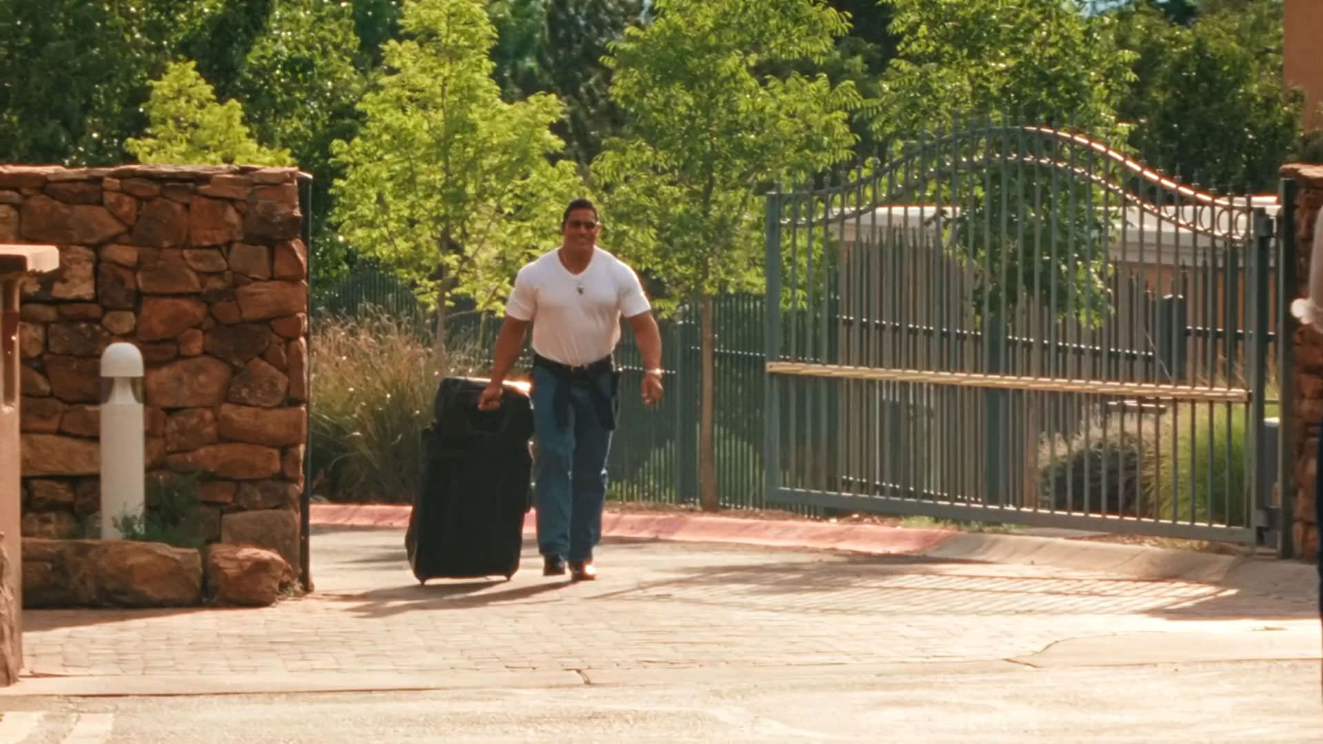 دواین جانسون راک با لباس سفید و چمدانی در دست از یک ساختمان خارج می‌شود در نمایی از فیلم ماشین کوبنده به کارگردانی بنی سفدی