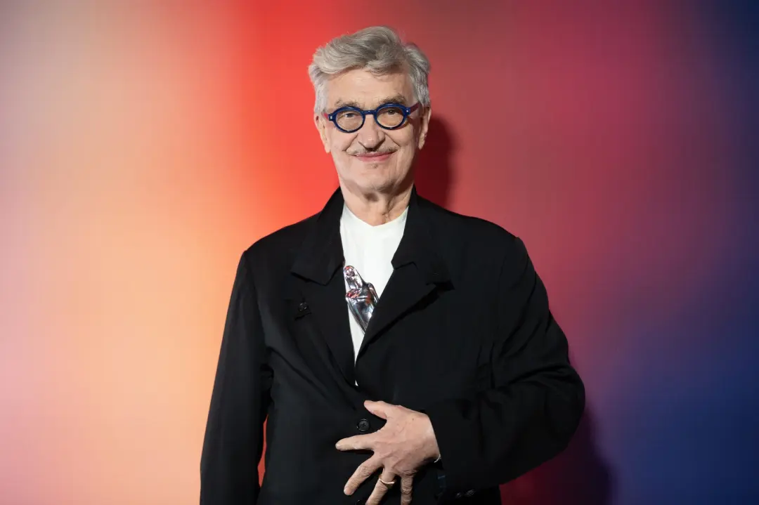 ویم وندرس (Wim Wenders) با عینک آبی و کت مشکی در مقابل پس‌زمینه رنگی